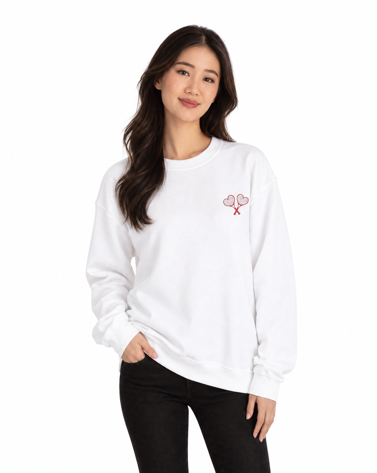 A woman modeling a white crewneck sweatshirt with a small red embroidered graphic of two hearts on the left chest, paired with black pants, against a plain white background.