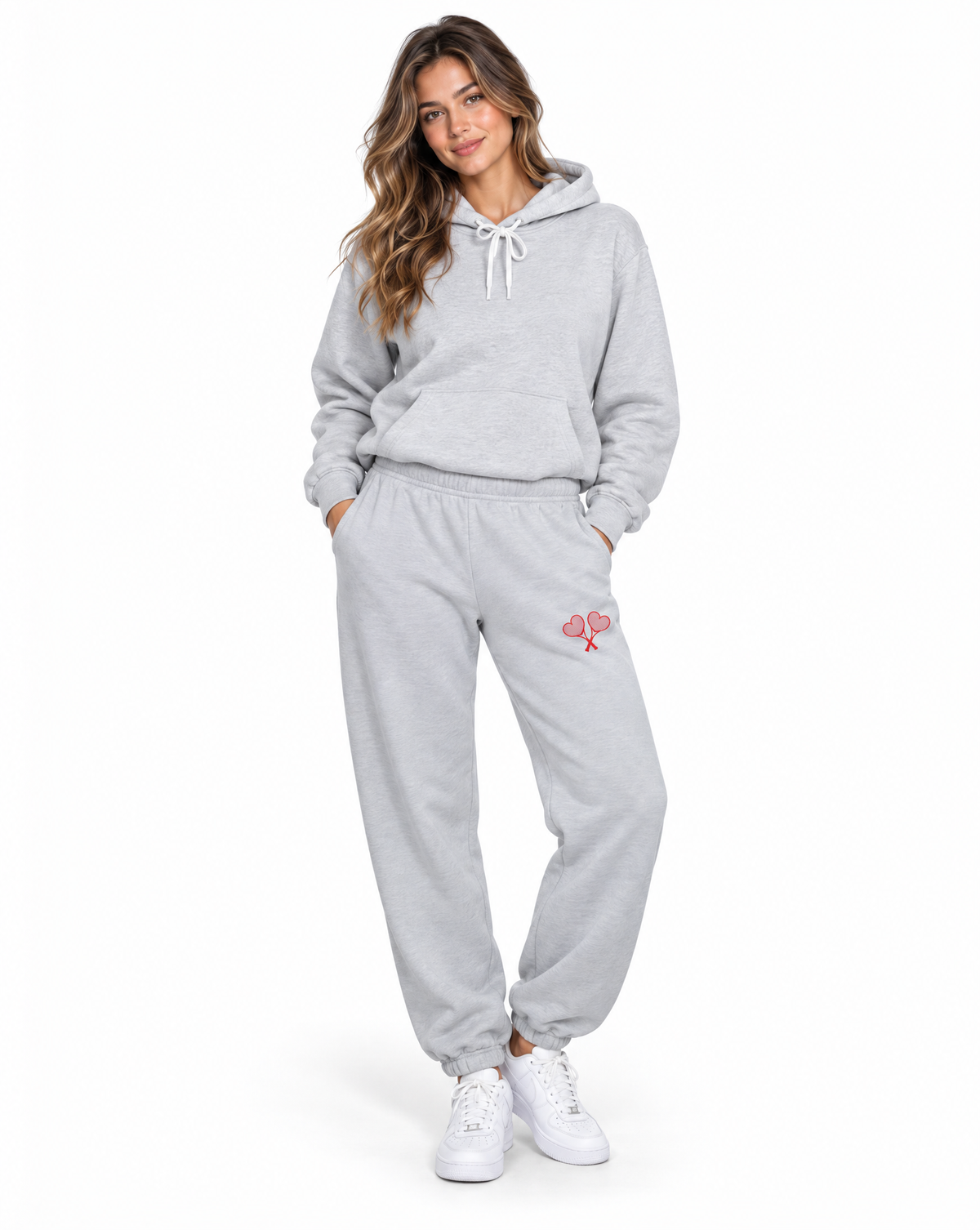 A woman modeling a light gray two-piece loungewear set consisting of a pullover hoodie and matching jogger pants, with a small pink graphic on the thigh, paired with white sneakers against a plain white background.