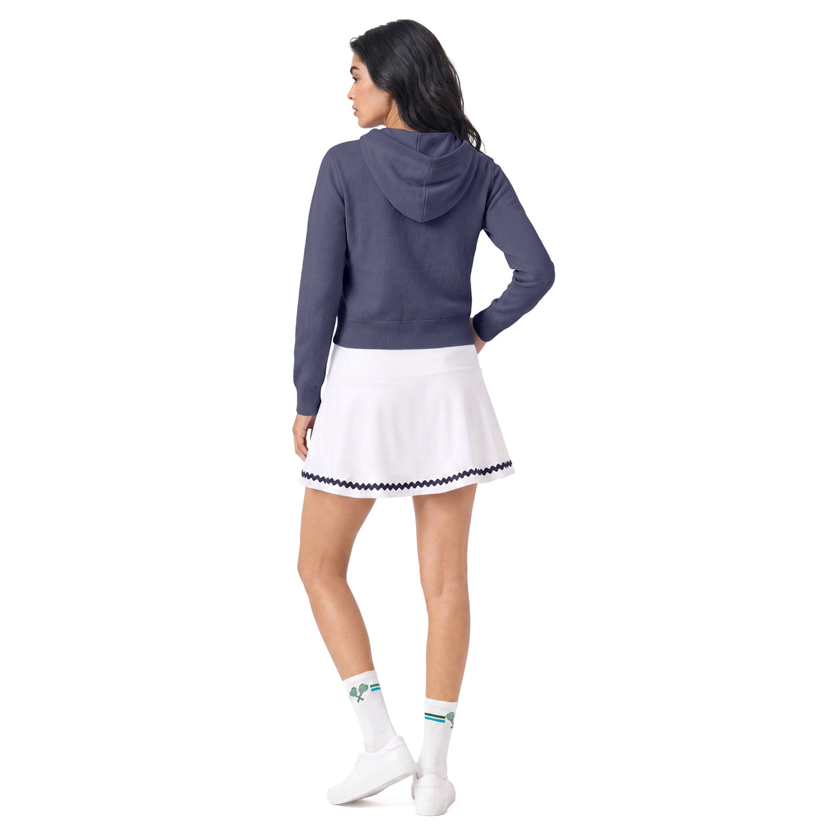 A woman is shown from the back wearing a slate blue cropped hoodie, a white tennis skirt with a navy and white checkered trim, white socks, and white sneakers, set against a plain white background.