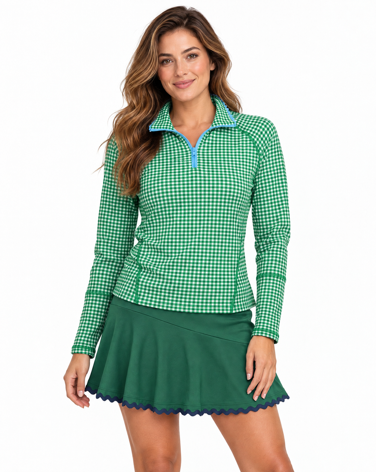 A woman modeling a green and white gingham long-sleeve quarter-zip pullover with light blue trim, paired with a dark green tennis skirt, against a solid white background.