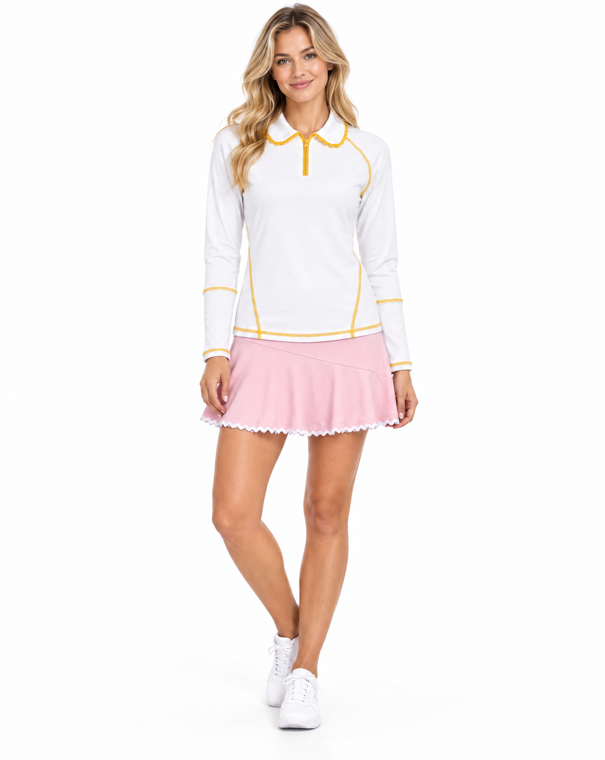 A woman modeling a white long-sleeve polo shirt with yellow trim and a light pink pleated tennis skirt, paired with white sneakers, against a plain white background.