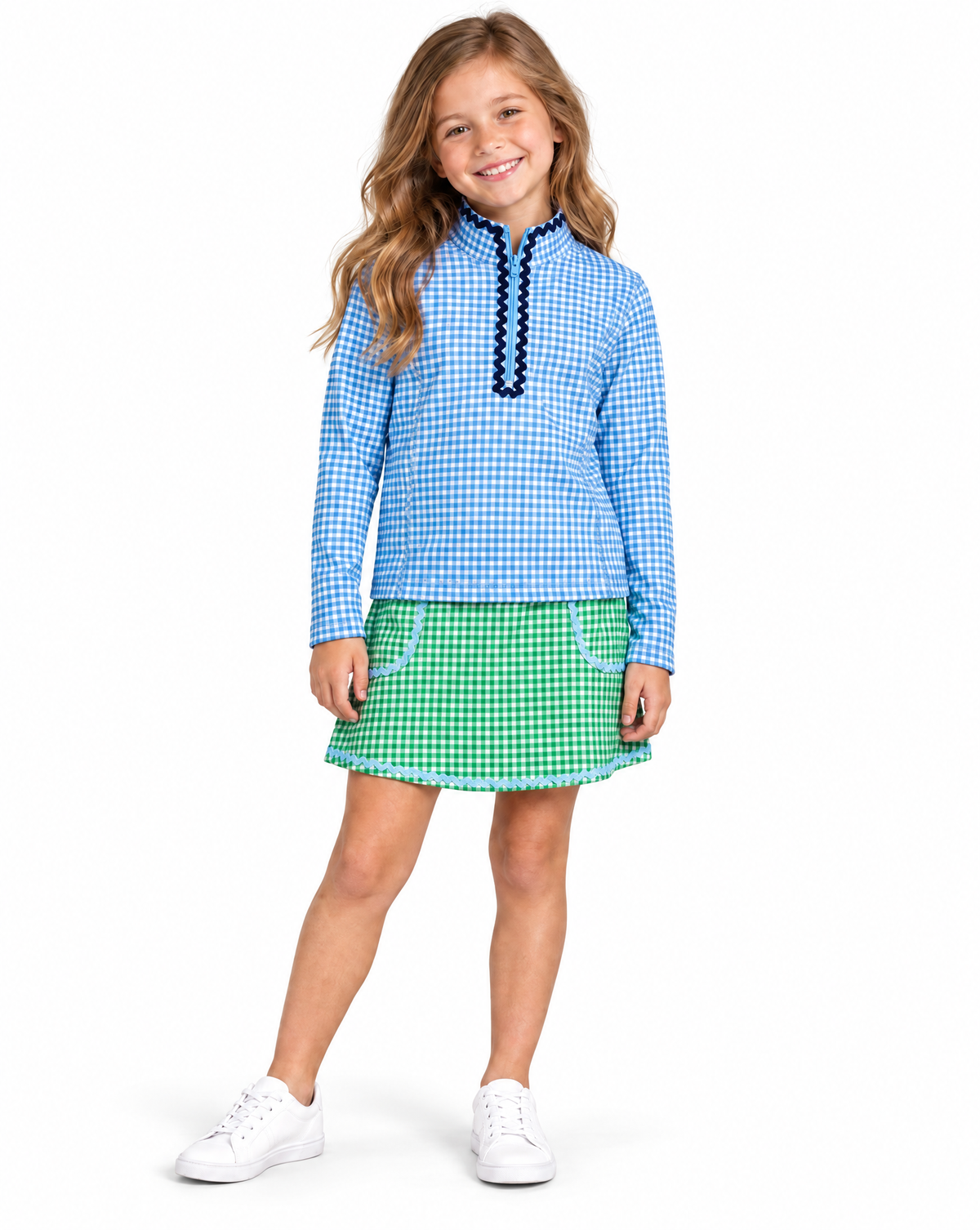 Young girl wearing a blue and white gingham golf top with navy ruffle trim and green and white gingham golf skirt with light blue trim on a white studio background