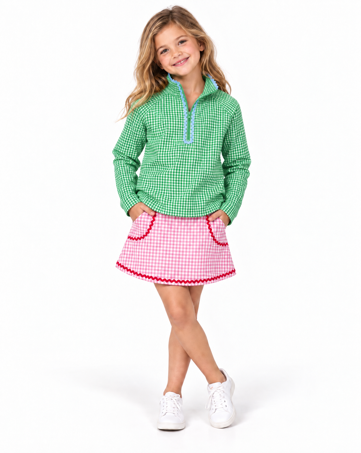 Young girl wearing a green and white gingham golf top with light blue ruffle trim and pink and white gingham golf skirt with red trim on a white studio background