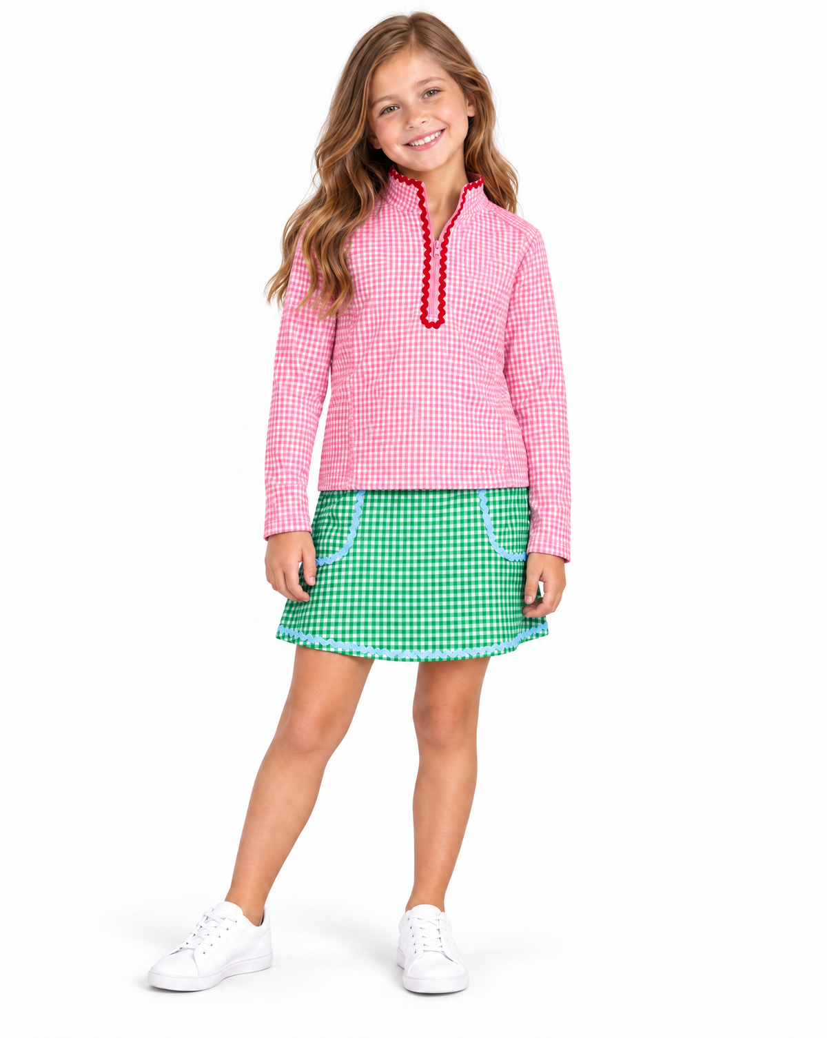 Young girl wearing a pink and white gingham golf top with red ruffle trim and green and white gingham golf skirt with light blue trim on a white studio background