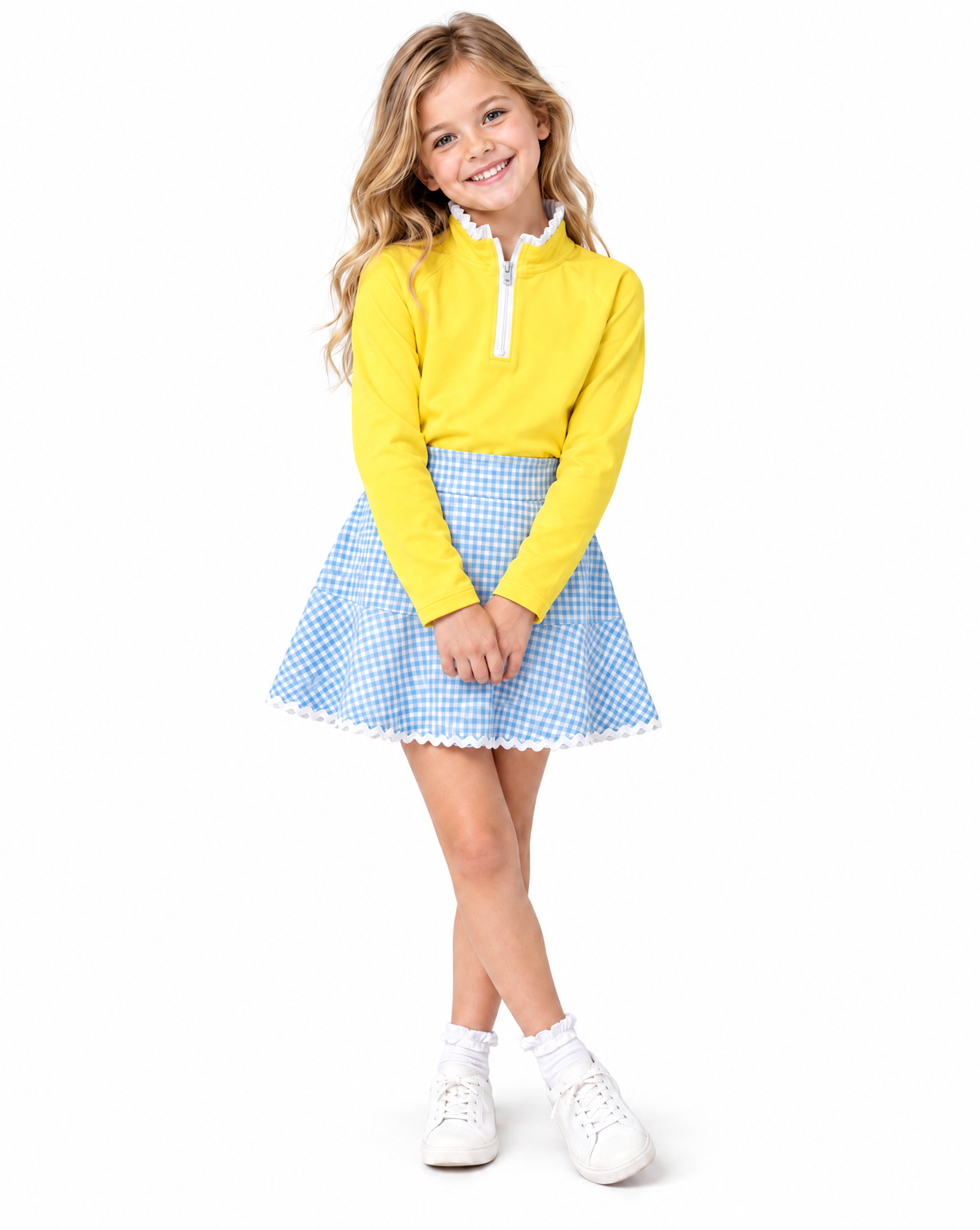 A young girl modeling a yellow long-sleeve half-zip pullover and a blue and white gingham pleated skirt, paired with white socks and white sneakers, against a solid white background.