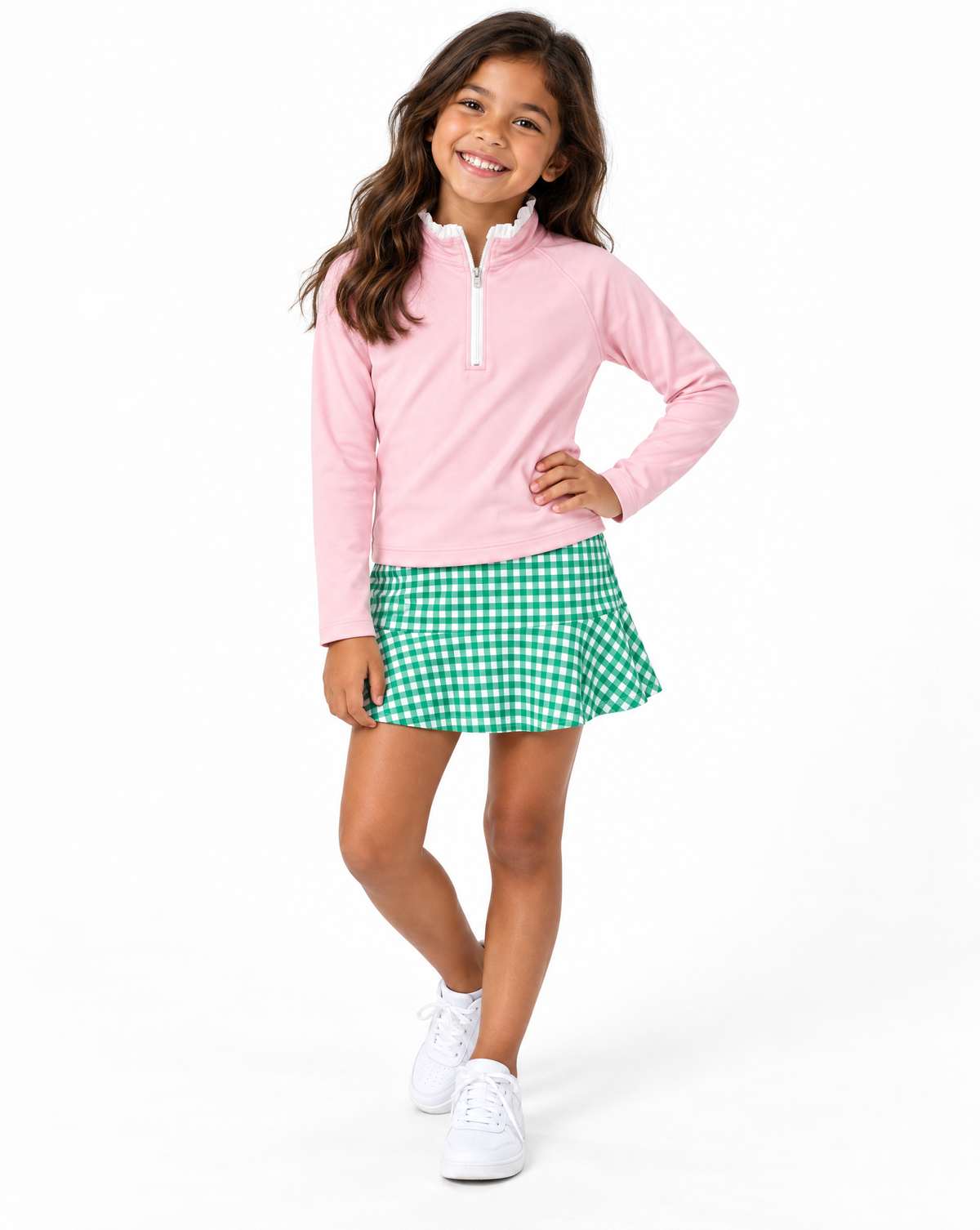 A young girl modeling a pink long-sleeve quarter-zip pullover and a green and white gingham mini skirt, paired with white sneakers, against a solid white studio background.