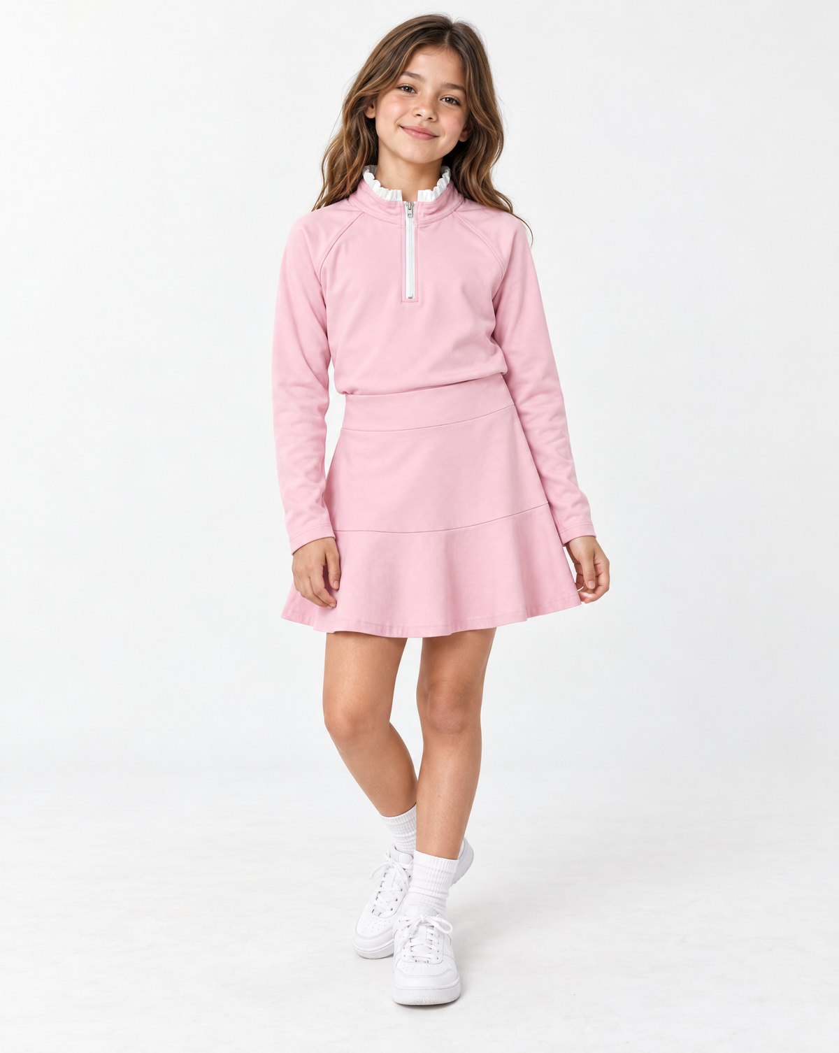 Young girl with blonde hair wearing a pink long sleeve athletic top with white trim and a pink tiered skirt and white sneakers against a flat white background