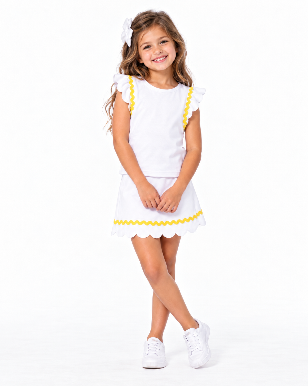 A young girl with long light brown hair wearing a white two-piece outfit with yellow scalloped trim, featuring a short-sleeve top with ruffle details and a matching skirt. She is also wearing a white hair bow and white sneakers, standing against a solid white studio background.