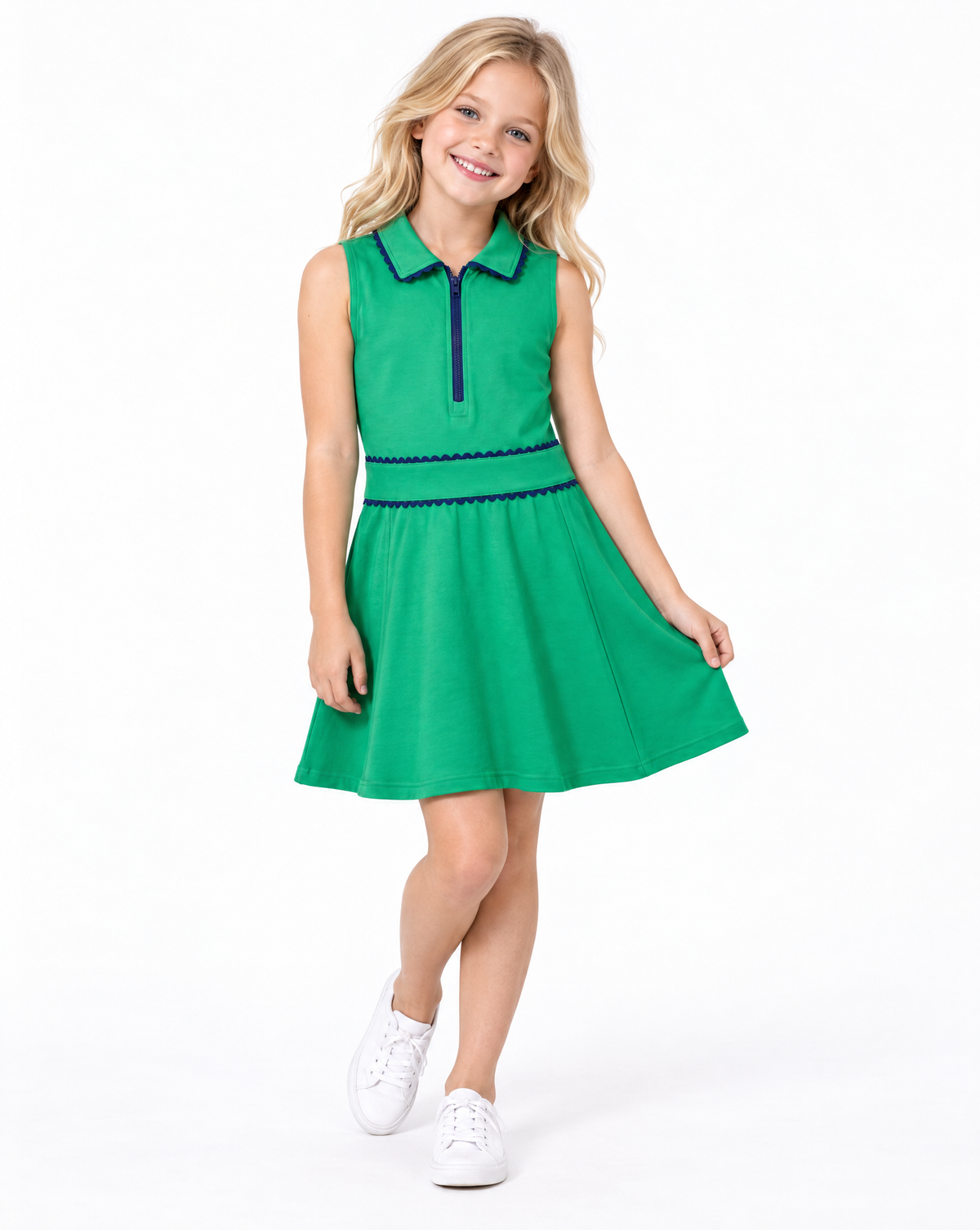 A young girl with blonde hair is modeling a bright green sleeveless polo dress with dark blue trim, paired with white sneakers, against a plain white studio background.