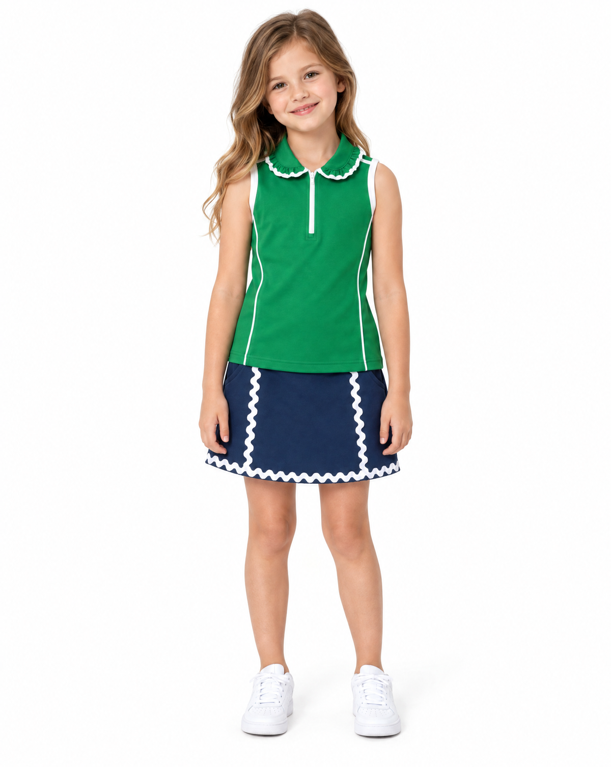 A young girl stands against a plain white background wearing a sleeveless green and navy blue top and skirt with white trim, paired with white sneakers.