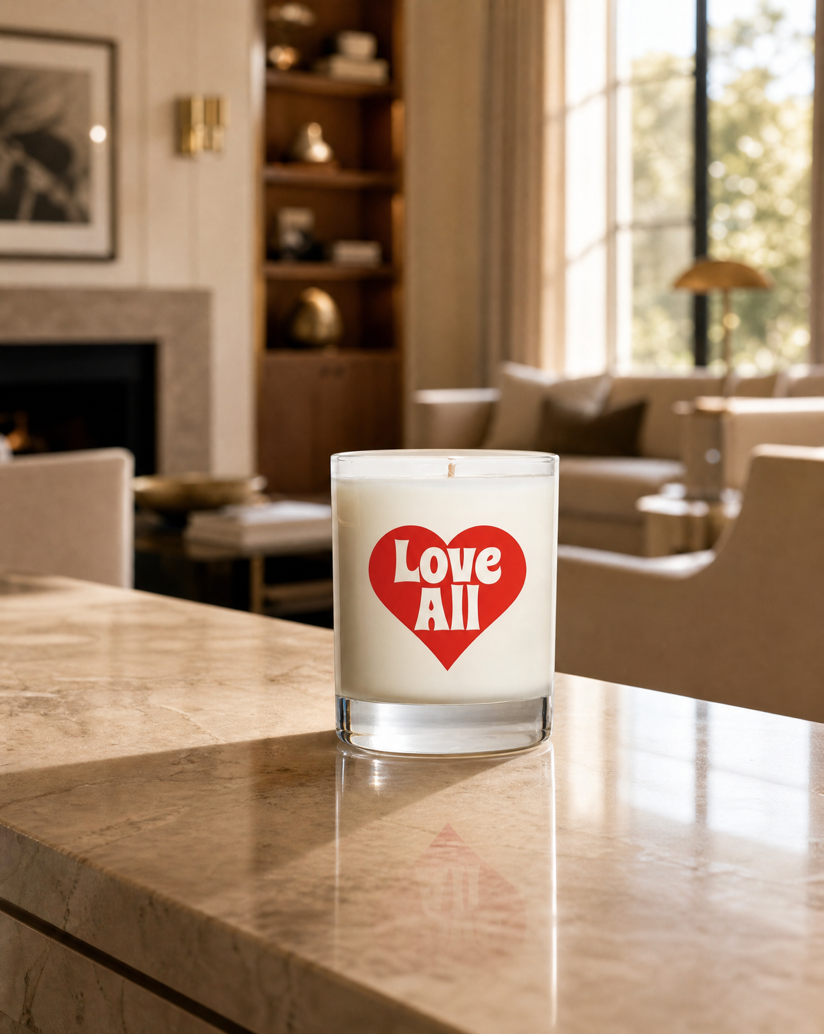 A white candle in a clear glass jar sits on a polished marble countertop in a warm, modern living room. The front of the candle features a red heart graphic with the words 'Love All' in white.