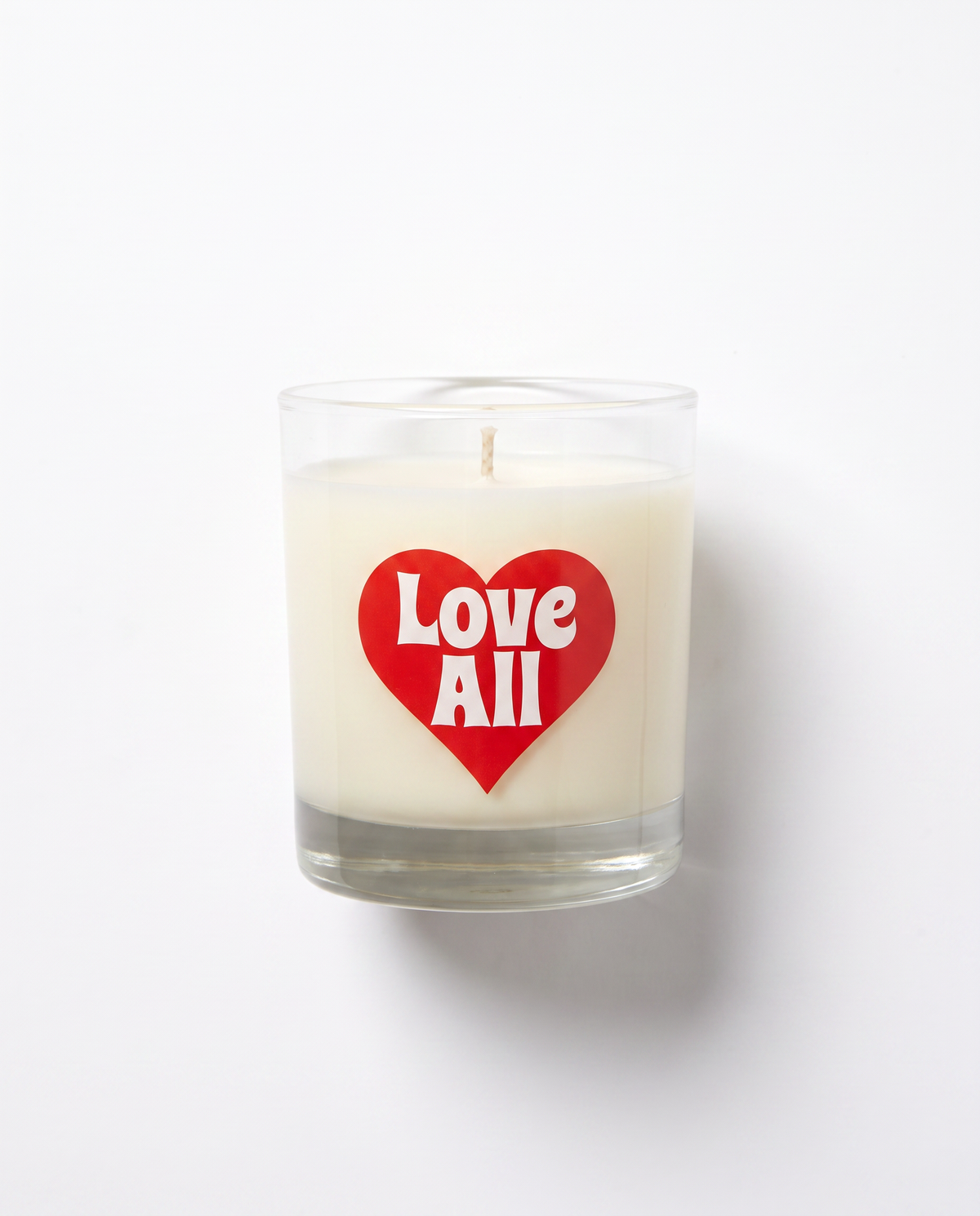 A white wax candle in a clear glass jar with a red heart-shaped sticker on the front that reads 'Love All' in white text, photographed against a plain white background.