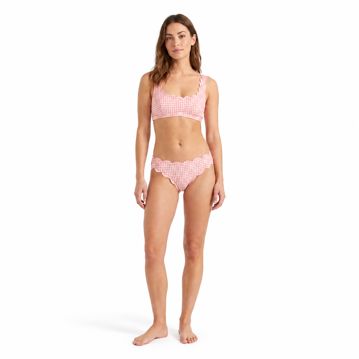 A woman modeling a two-piece bikini with a pink and white gingham pattern and scalloped edges, standing against a solid white background.