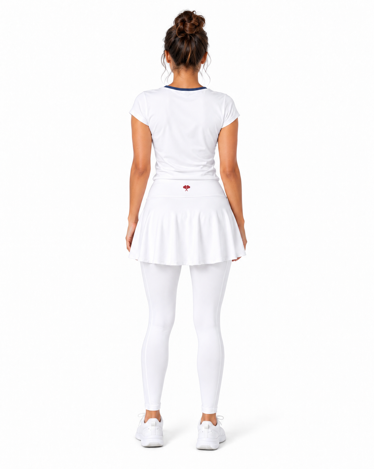 A woman viewed from the back wearing a white tennis outfit consisting of a short-sleeve top with a thin dark blue collar, a pleated tennis skirt with a small red heart logo on the back, white leggings, and white sneakers, set against a plain white background.