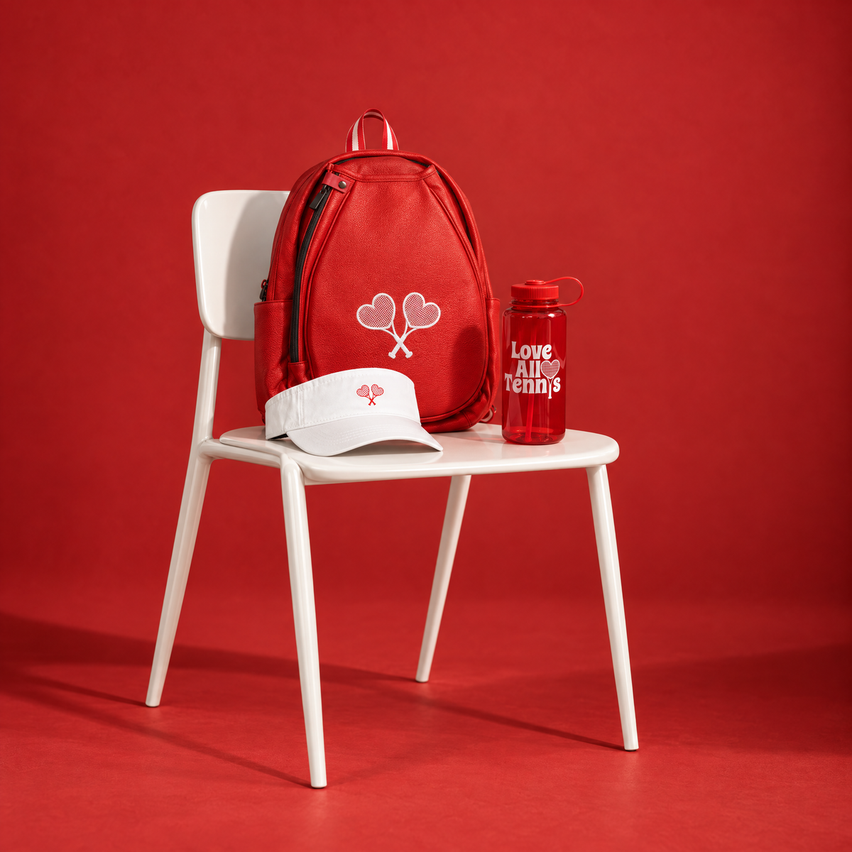 A red backpack with a white heart-and-shuttlecock logo sits on a white chair, styled with a matching red water bottle labeled 'Love Alice Tennis' and a white visor with a small red logo, all against a solid red studio background.