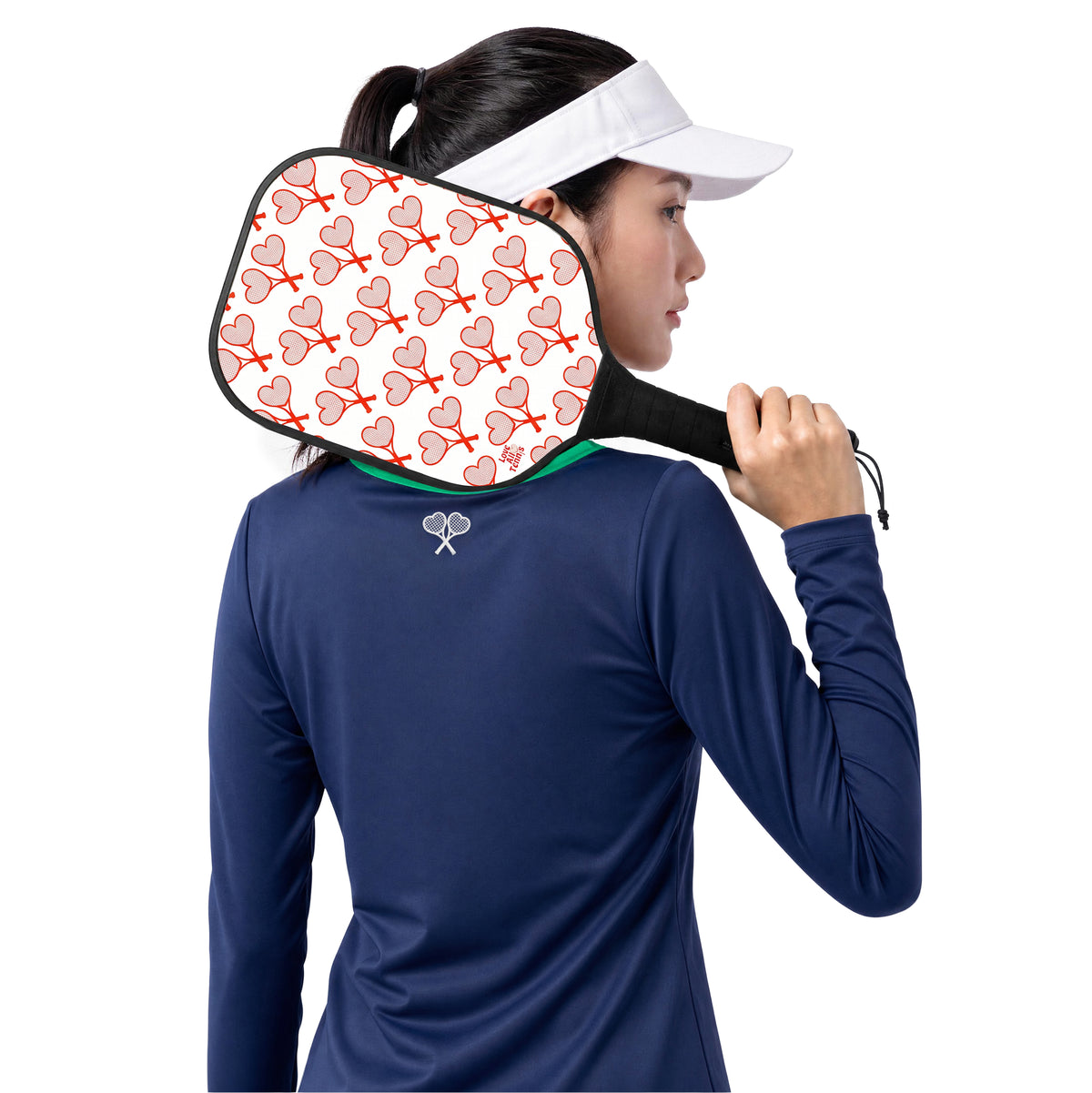 A woman in a navy long-sleeve athletic shirt and white visor holds a pickleball paddle against her neck. The paddle has a white face with a repeating red and pink heart pattern and black trim.