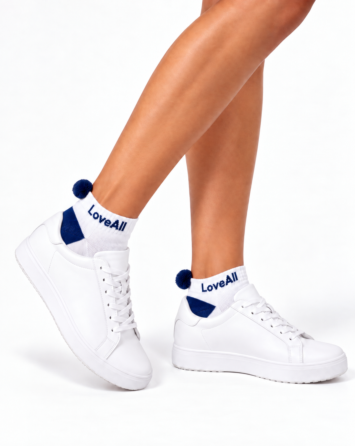 White low-top sneakers worn with white and navy pom pom socks printed with 'LoveAll', shown on a model's feet against a white background.