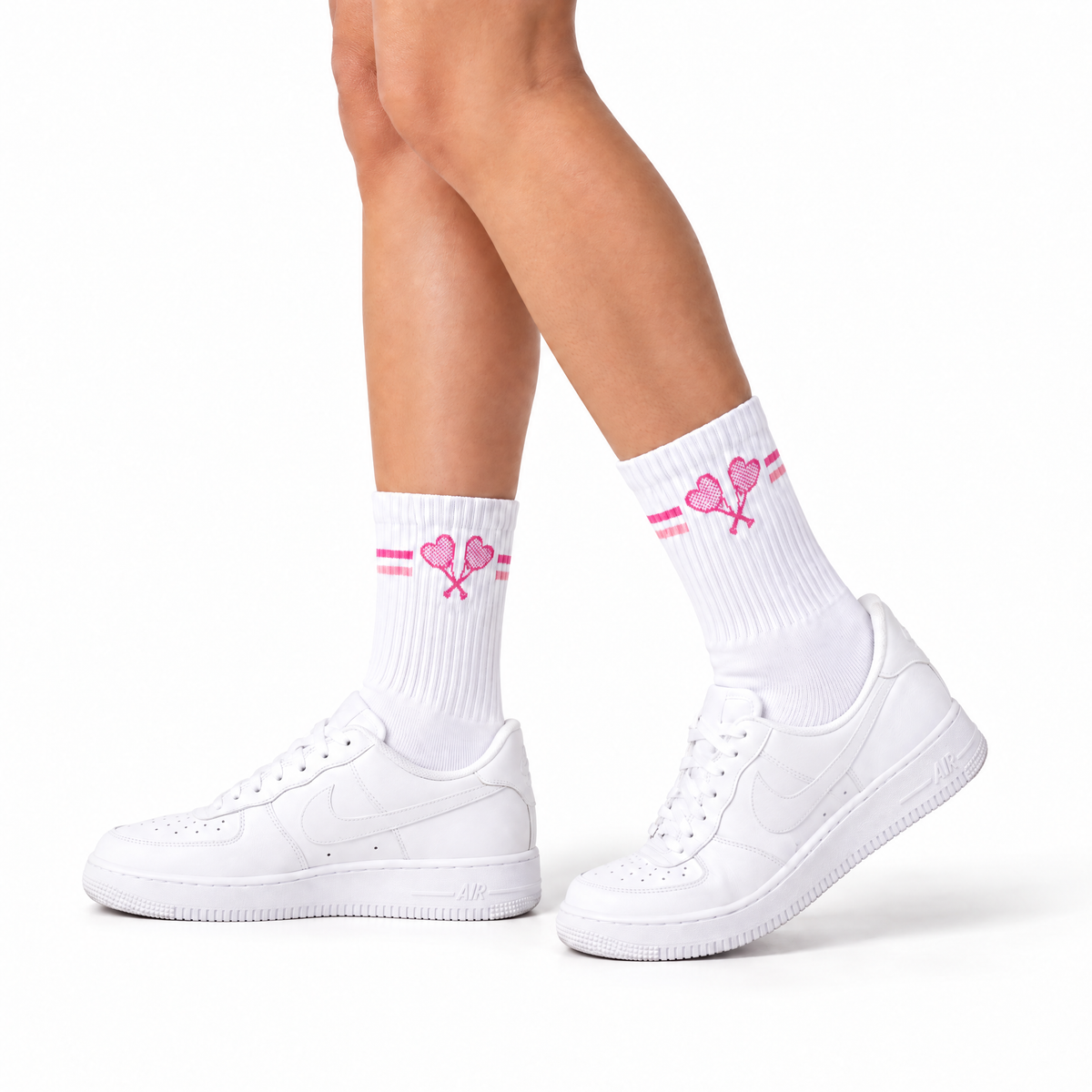 White crew socks with pink heart and tennis racket graphics, worn with white low-top sneakers on a model's legs against a white background.