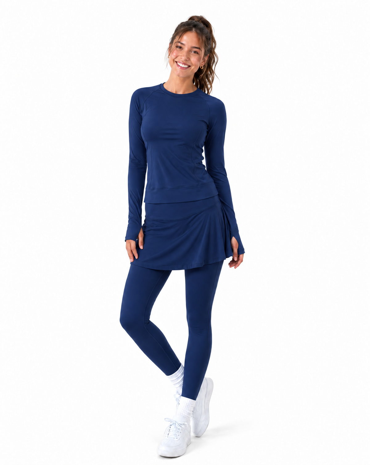 Woman wearing navy athletic long sleeve top and skirted leggings and sneakers set against a flat white background