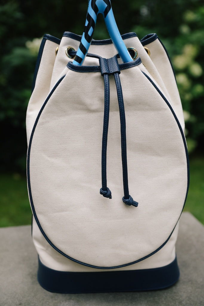 An off-white canvas bucket bag with navy blue trim, drawstring closure, and a blue-and-white striped top handle, photographed outdoors on a stone surface with blurred green foliage in the background.