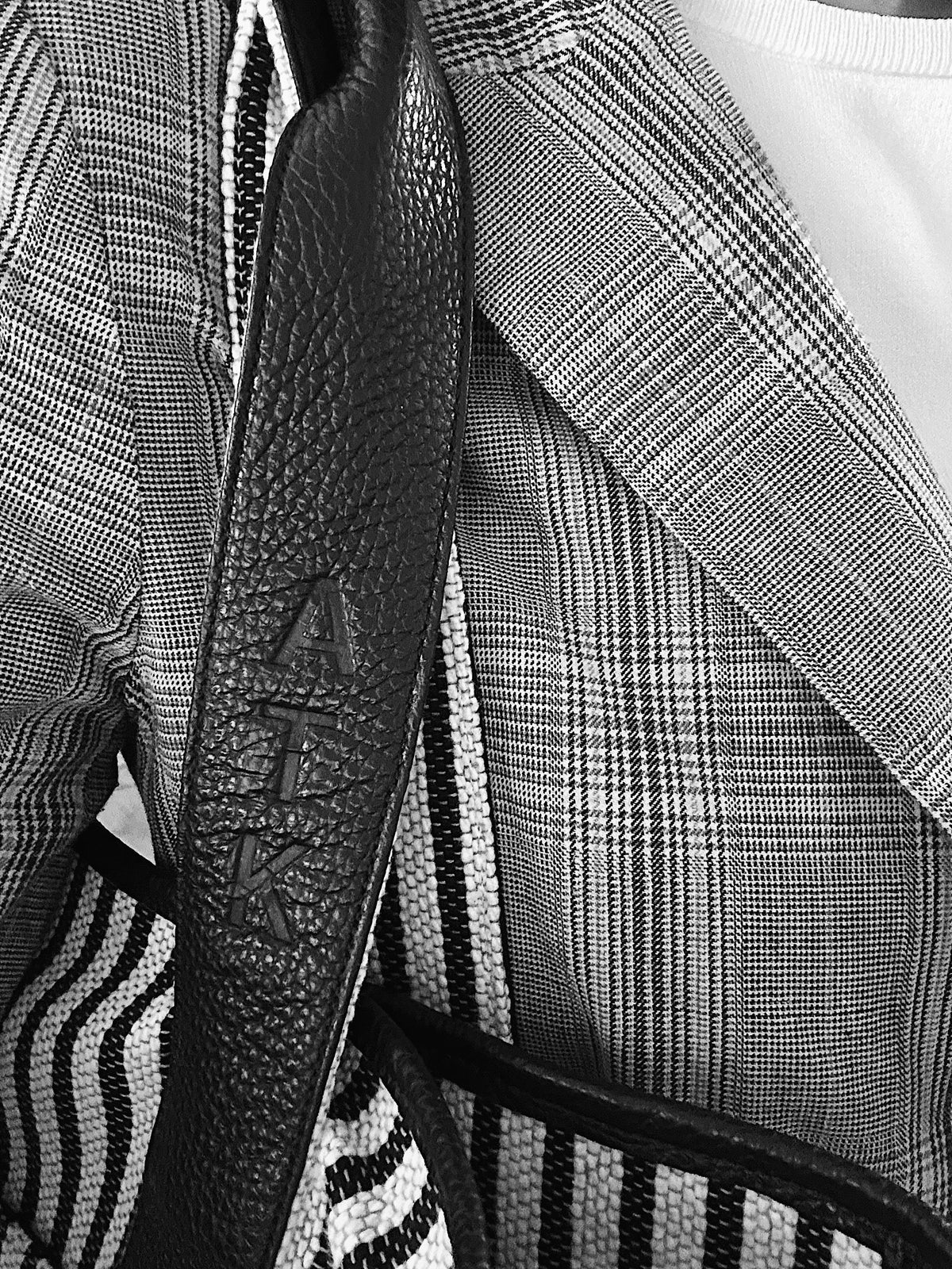 Close-up of a black textured leather shoulder strap with the brand name 'ATK' embossed vertically, worn over a black-and-white plaid blazer and striped garment.