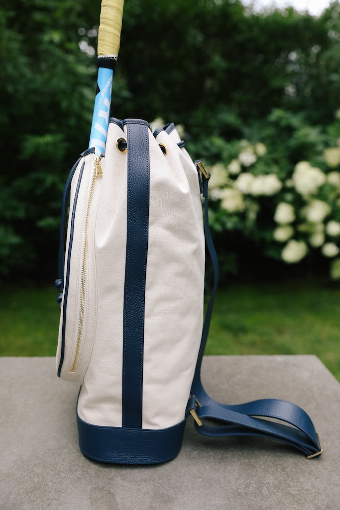 A canvas golf bag with navy blue trim and a blue-and-white striped grip visible on the club handle, set outdoors on a stone surface with a blurred garden background.