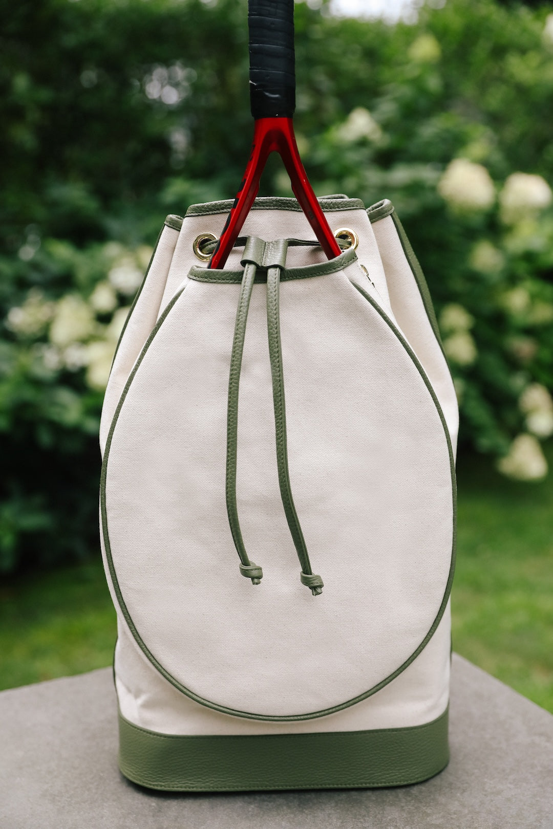 A canvas drawstring bucket bag in off-white and olive green, featuring a large oval front pocket and a black-and-red hanging strap, set against a blurred garden background.