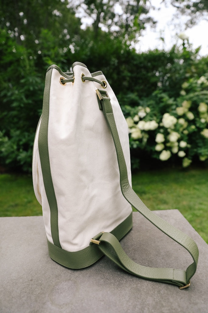 An off-white canvas bucket bag with olive green leather trim and strap, resting on a gray stone surface with a blurred garden background.
