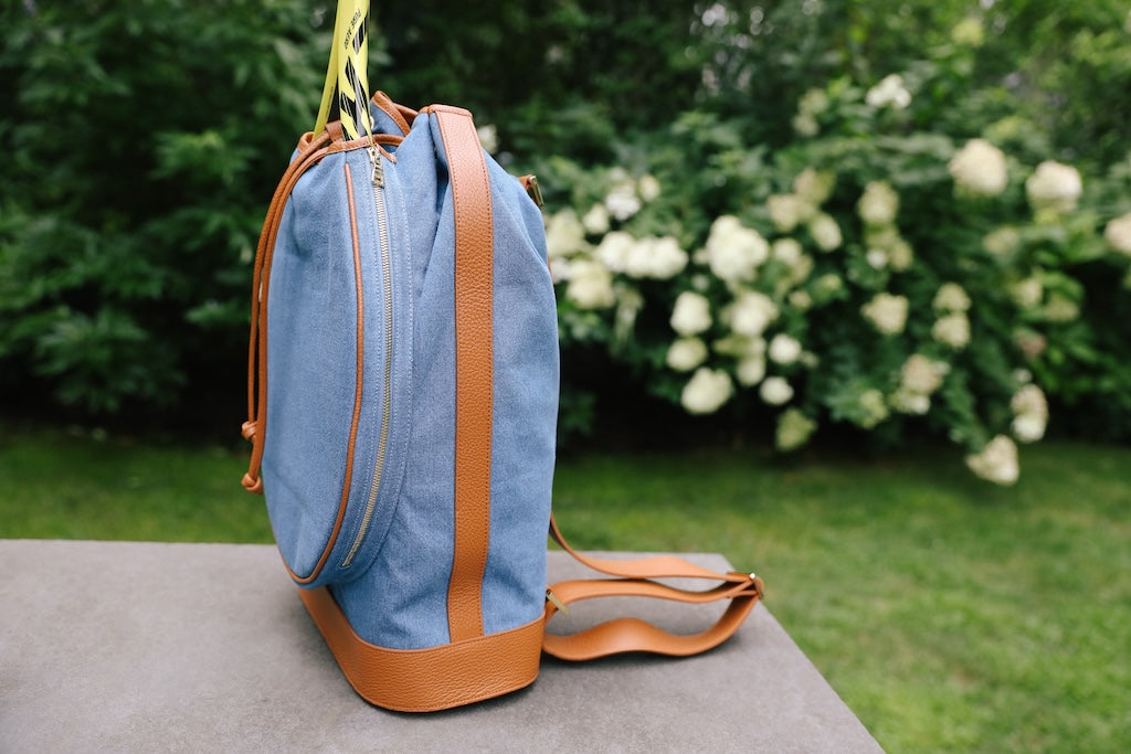A light blue denim backpack with tan leather trim and straps, standing on a stone surface with a blurred garden background of green foliage and white flowers.