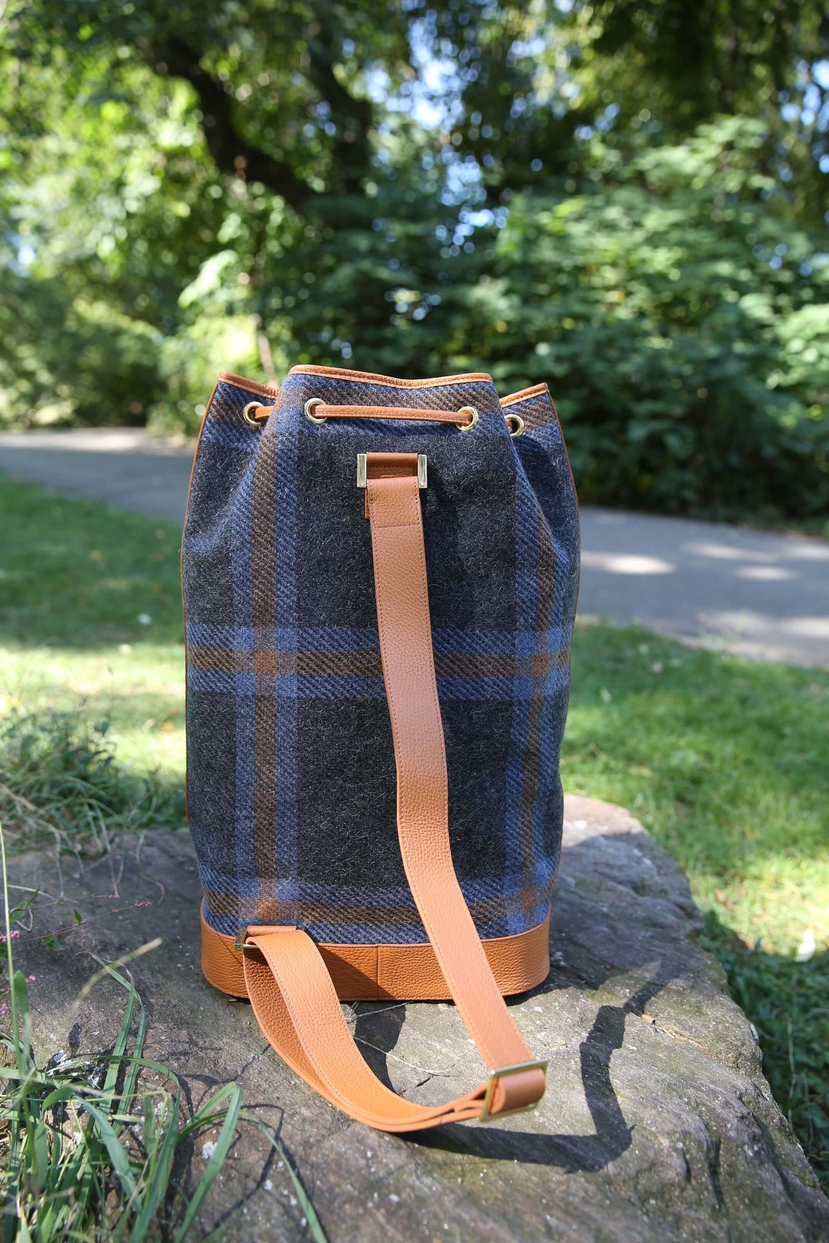 A plaid bucket bag made of dark blue and brown wool, with tan leather trim and strap, sitting on a stone in a sunny park setting.