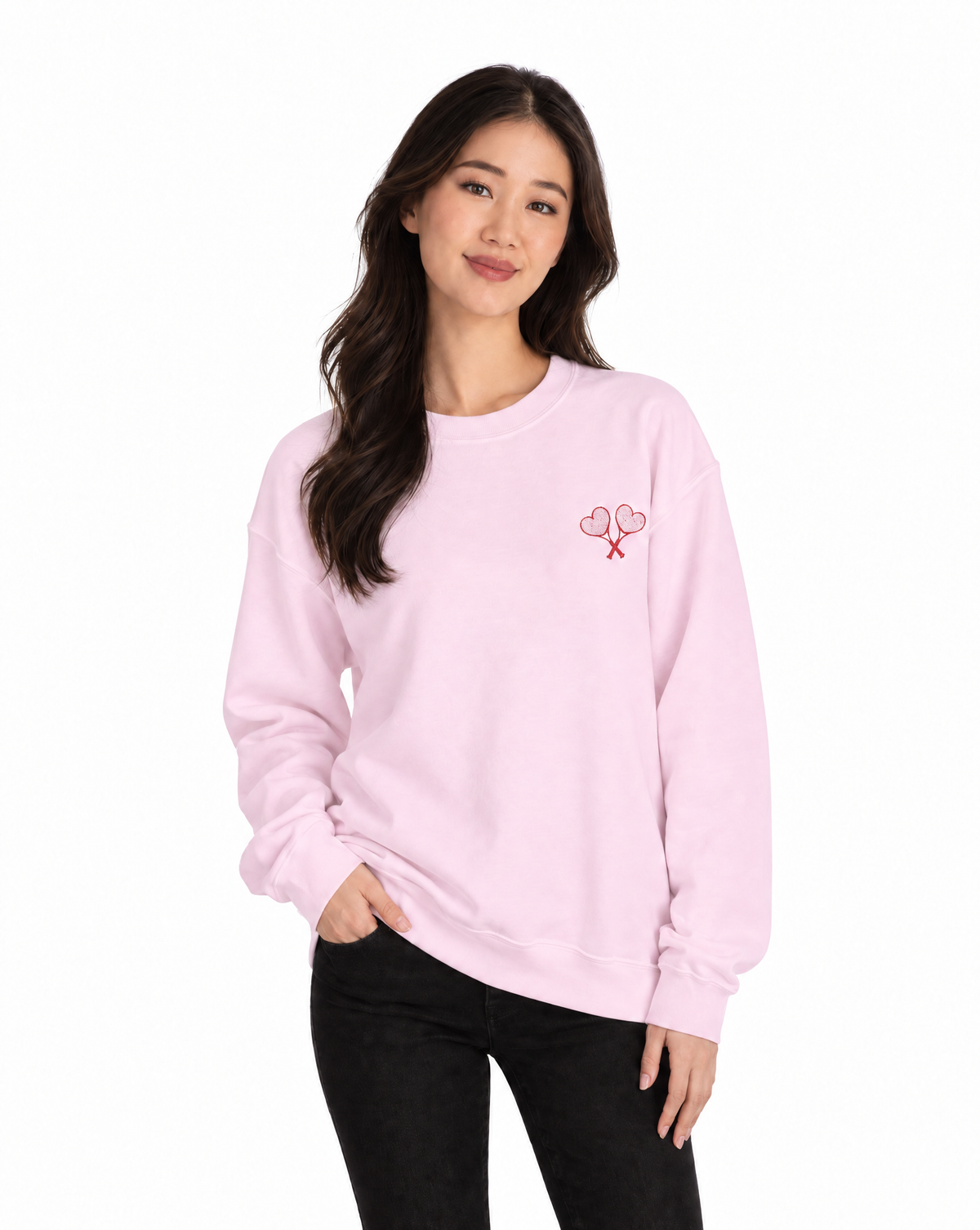 A woman modeling a light pink crewneck sweatshirt with a small red embroidered graphic of two hearts and crossed keys on the left chest, paired with black pants against a plain white background.
