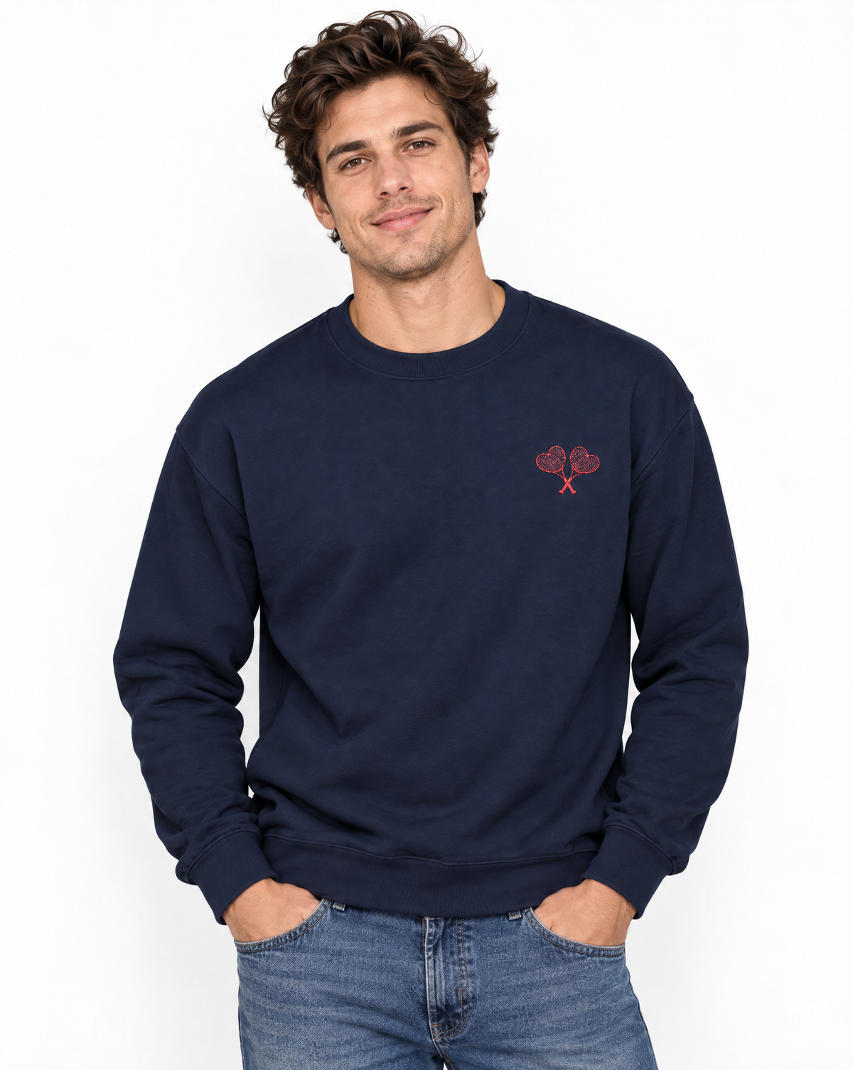 A man wearing a navy blue crewneck sweatshirt with a small red embroidered heart and crossed keys motif on the left chest, paired with blue jeans, against a plain white background.