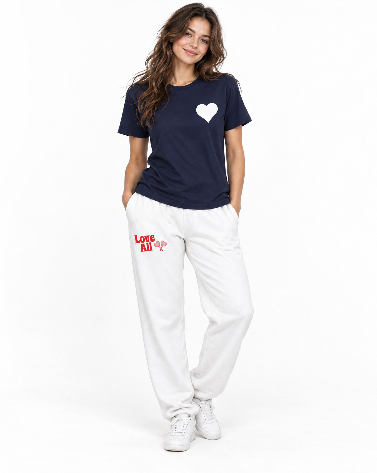 A woman modeling a navy blue t-shirt with a white heart on the chest and white sweatpants with red 'LOVE ALL' text on the thigh, paired with white sneakers, against a plain white background.