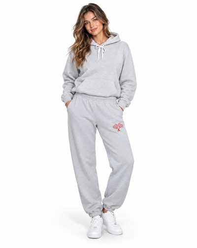 A woman modeling a light gray two-piece loungewear set consisting of a pullover hoodie and matching jogger pants, with a small pink graphic on the thigh, paired with white sneakers against a plain white background.