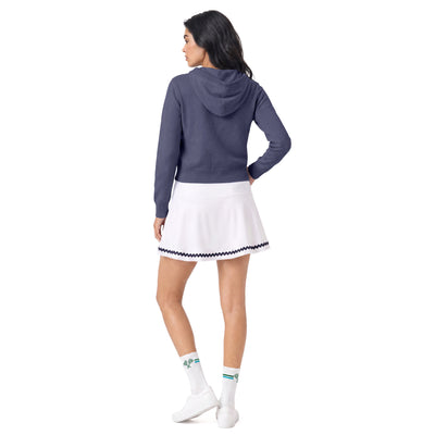 A woman is shown from the back wearing a slate blue cropped hoodie, a white tennis skirt with a navy and white checkered trim, white socks, and white sneakers, set against a plain white background.