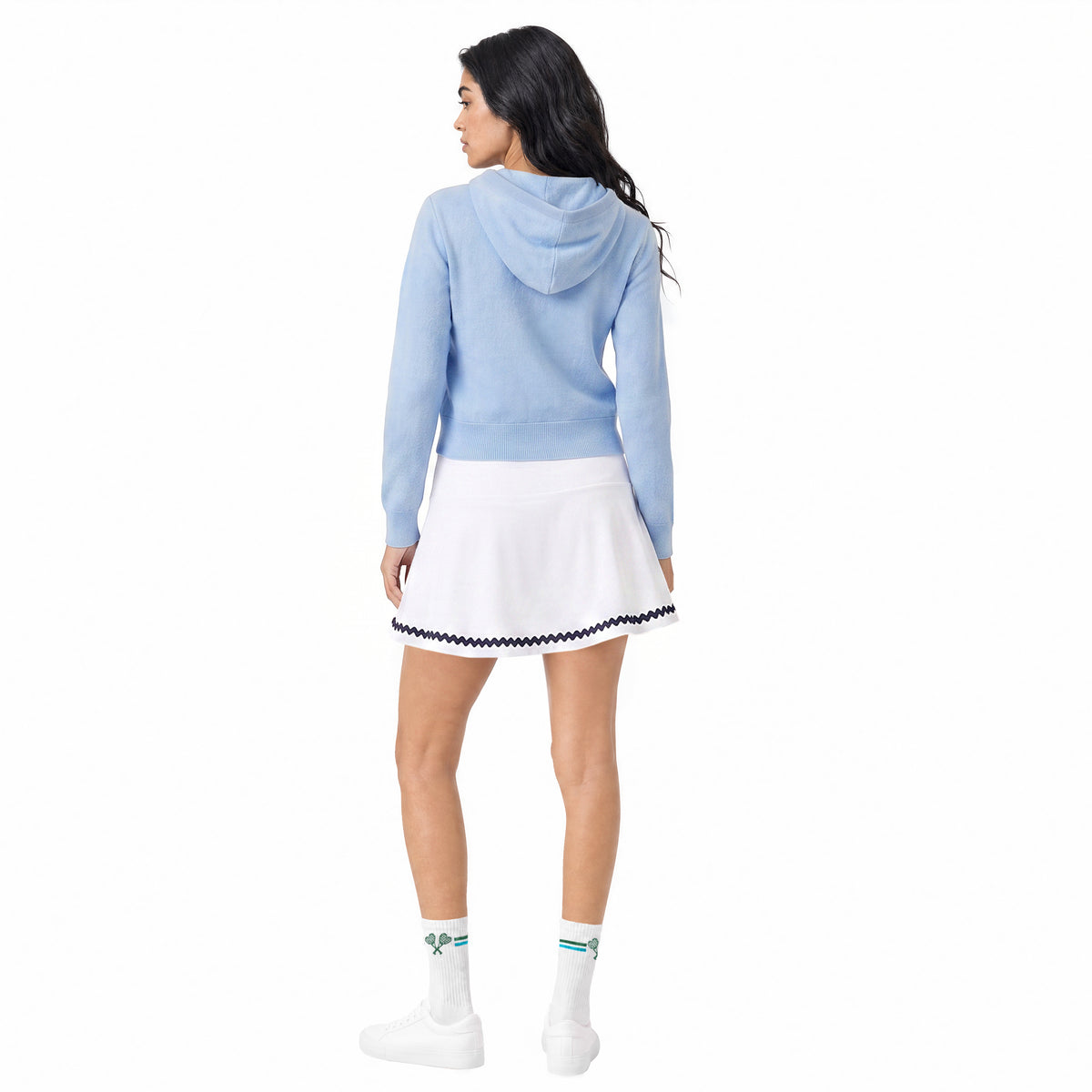 A woman shown from the back wearing a light blue hooded sweatshirt, a white tennis skirt with navy trim, white socks, and white sneakers against a plain white background.