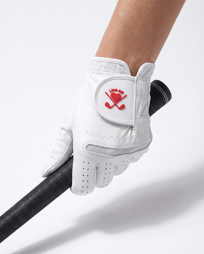 A white golf glove with a red logo featuring a heart and crossed clubs, worn on a hand holding a black golf club shaft against a white background.