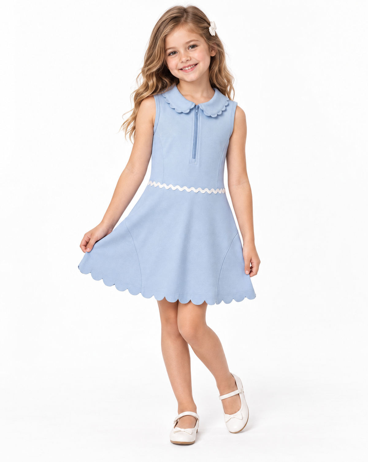 A young girl with blonde hair wearing a light blue sleeveless dress with a white scalloped collar, waistband, and hem, paired with white Mary Jane shoes, standing against a white studio background.