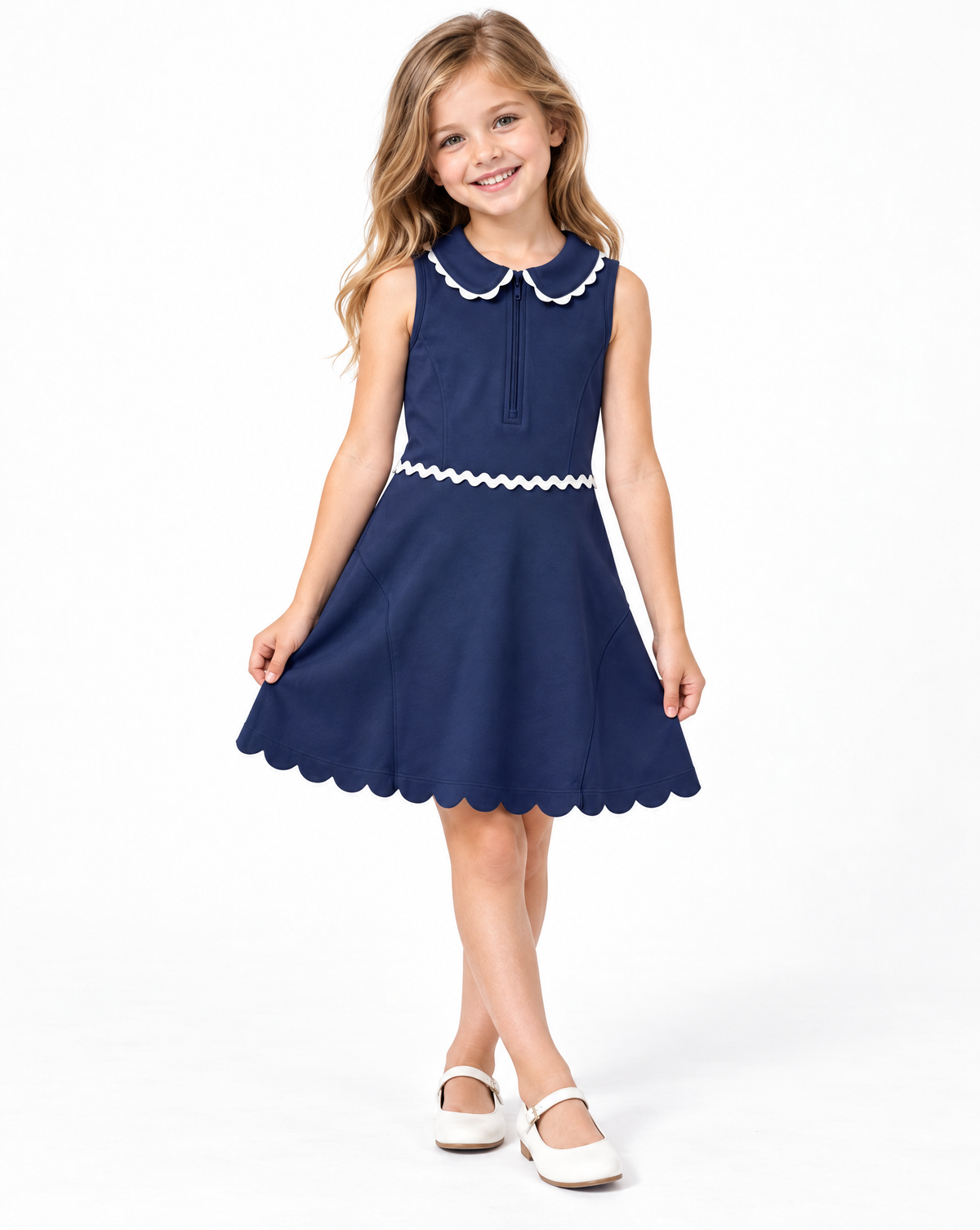 A young girl with blonde hair smiling and wearing a sleeveless navy blue dress with a white scalloped Peter Pan collar, a matching waistband, and white Mary Jane shoes, photographed against a seamless white background.