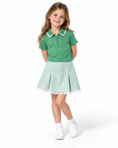 A young girl modeling a green short-sleeve polo shirt with a white ruffled collar and a light green pleated skirt with a white ruffled hem, paired with white socks and sneakers, on a solid white background.