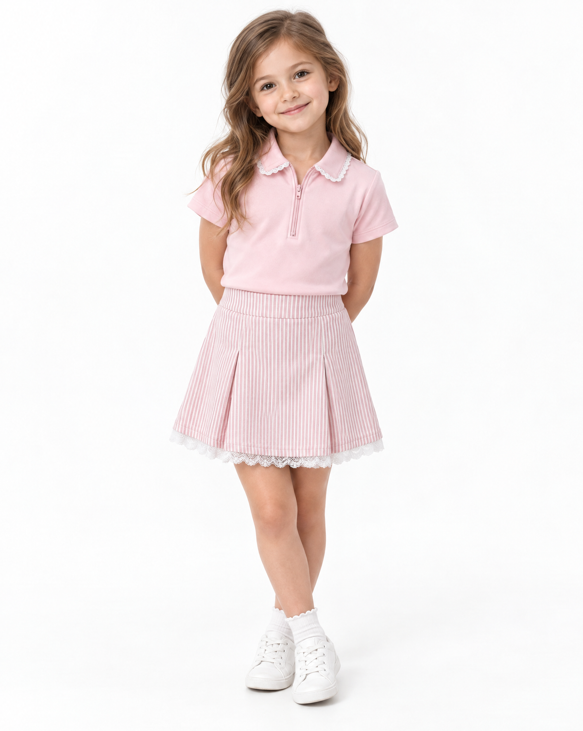 A young girl modeling a light pink short-sleeve polo shirt and a pink and white vertically striped pleated skirt with a white lace hem, paired with white sneakers, against a plain white studio background.
