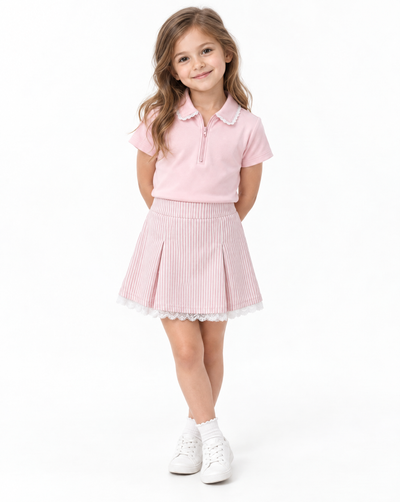 A young girl modeling a light pink short-sleeve polo shirt and a pink and white vertically striped pleated skirt with a white lace hem, paired with white sneakers, against a plain white studio background.