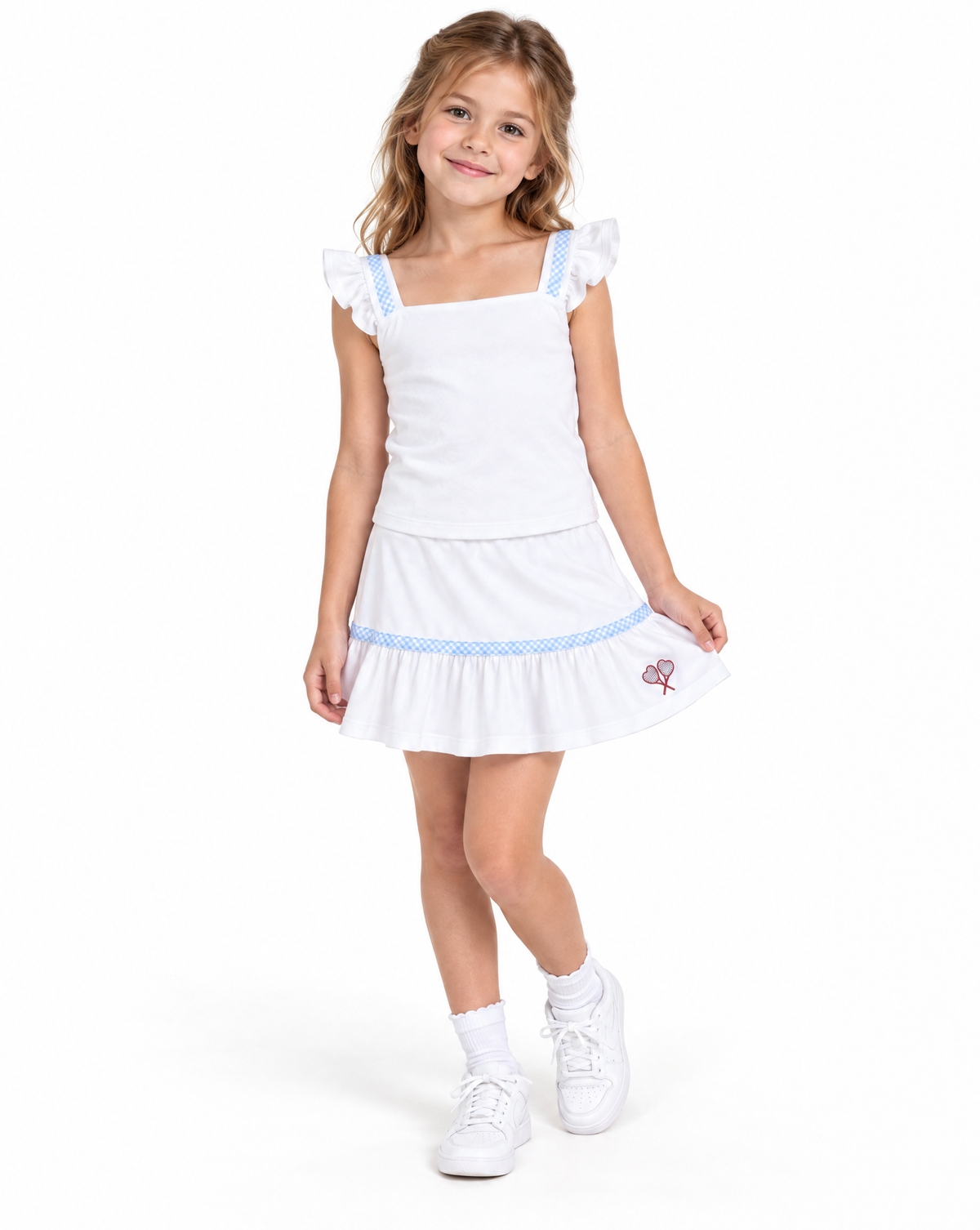 A young girl modeling a white sleeveless top and skirt with ruffled shoulder straps, a square neckline, and a ruffled hem trimmed with light blue polka dots. She is wearing white socks and white sneakers against a solid white studio background.