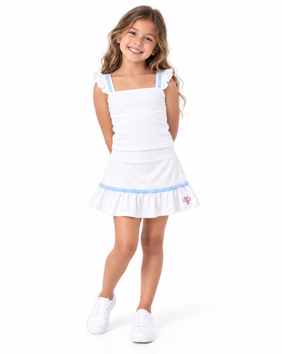 A young girl with long brown hair stands against a white background wearing a white sleeveless top and skirt with light blue trim and white sneakers.