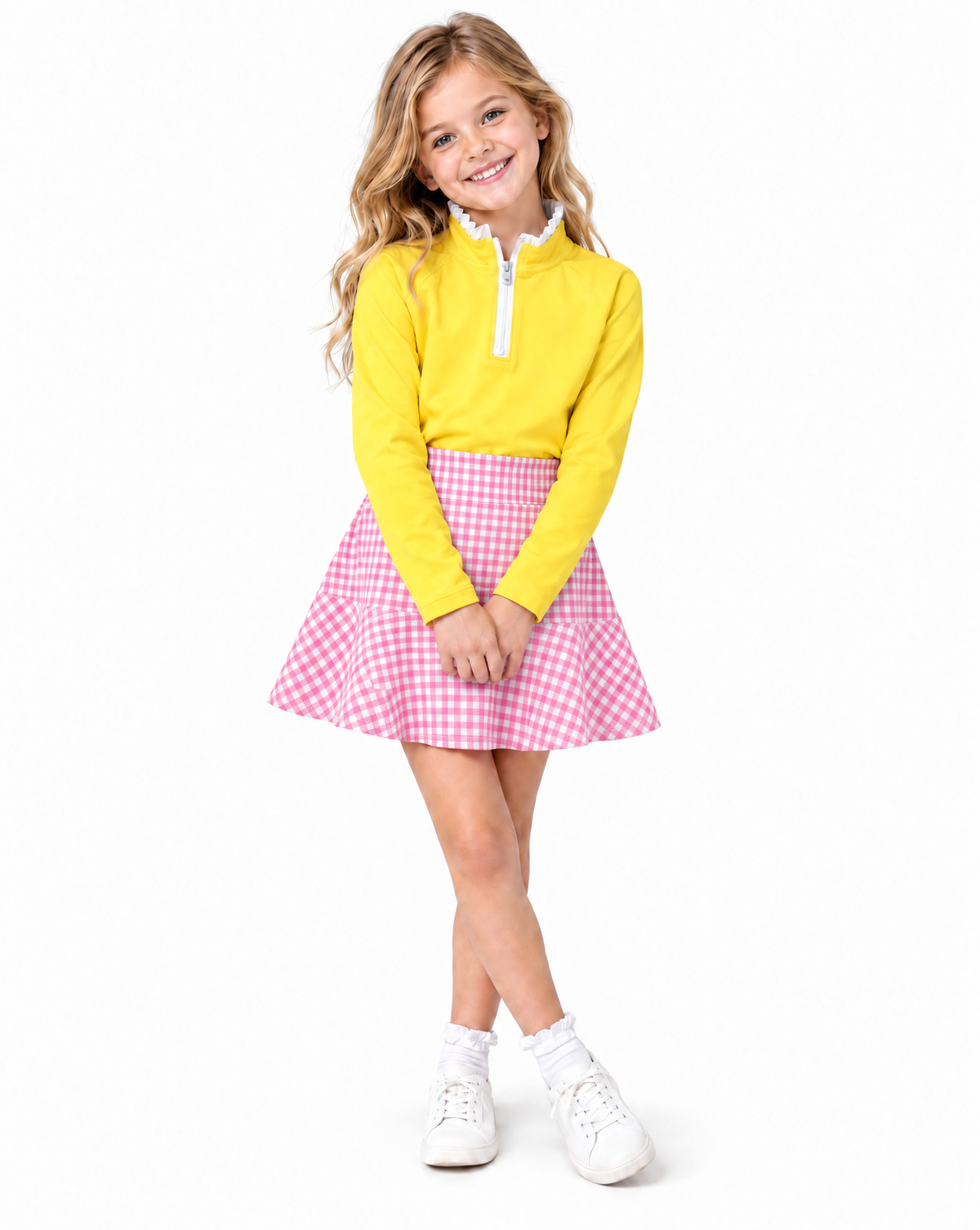A young girl with blonde hair stands against a white background wearing a bright yellow long-sleeve quarter-zip pullover, a pink and white gingham pleated skirt, white socks, and white sneakers.