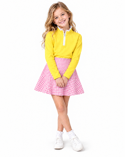 A young girl with blonde hair stands against a white background wearing a bright yellow long-sleeve quarter-zip pullover, a pink and white gingham pleated skirt, white socks, and white sneakers.