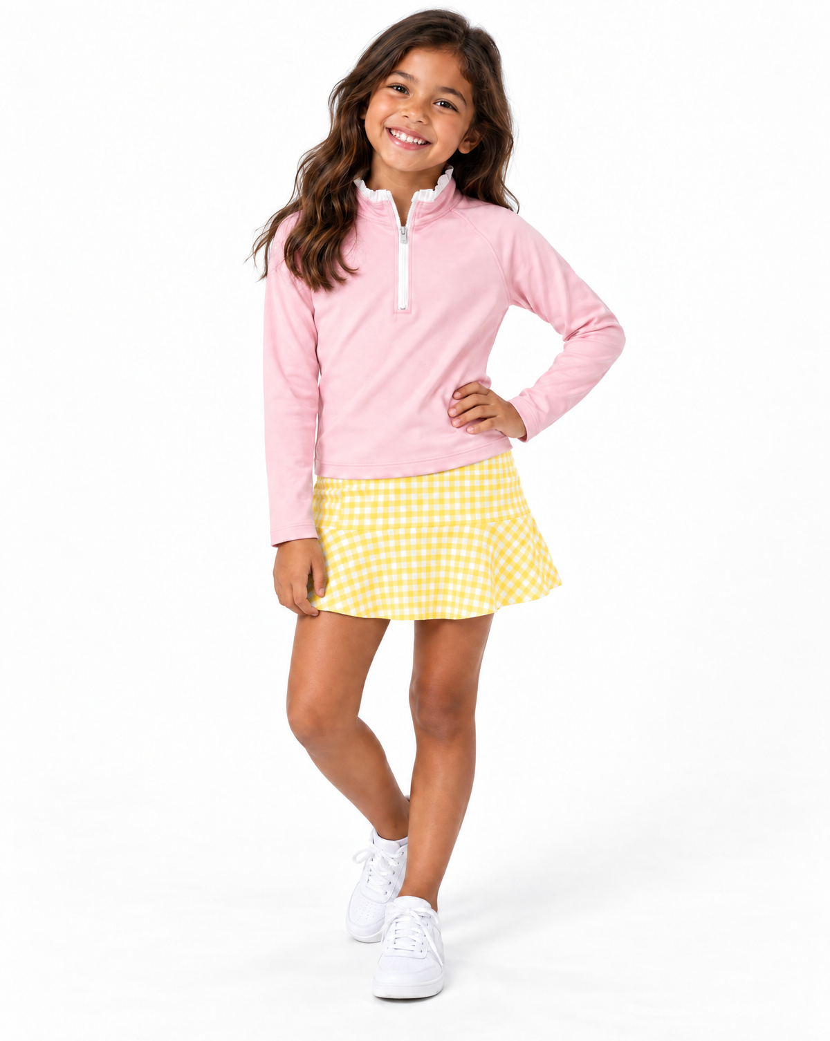 A young girl modeling a light pink long-sleeve quarter-zip pullover and a yellow and white gingham mini skirt, paired with white sneakers, against a solid white studio background.