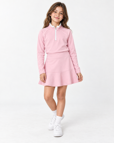 Young girl with blonde hair wearing a pink long sleeve athletic top with white trim and a pink tiered skirt and white sneakers against a flat white background