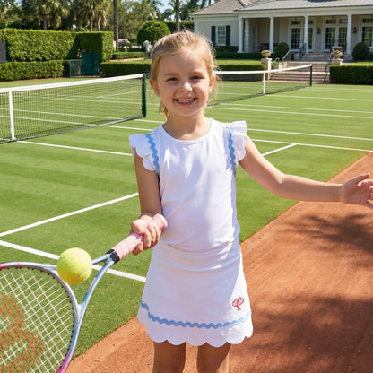 Young girl on a tennis court holding a racket and ball, with a house and palm trees in the background.