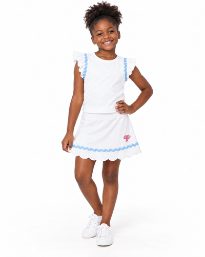 A young girl modeling a white two-piece outfit consisting of a short-sleeve top and a matching flared skirt, both decorated with light blue scalloped trim. The skirt features a small pink embroidered flower on the lower left side.